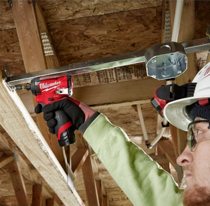 Milwaukee M12 Fuel 1/4" Impact Driver held by worker wearing gloves and safety glasses, red and black compact tool.