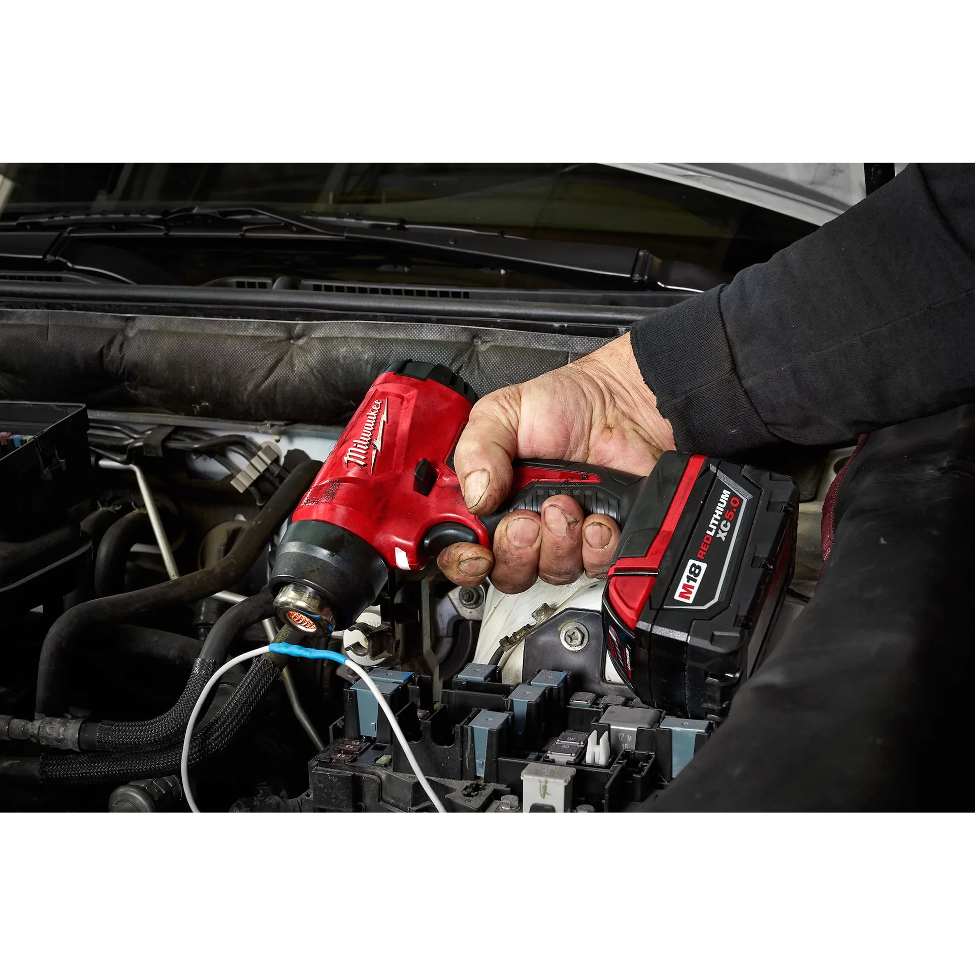 Milwaukee M18 Heat Gun 2688-20 red cordless tool held over engine bay with black battery pack and ergonomic handle.